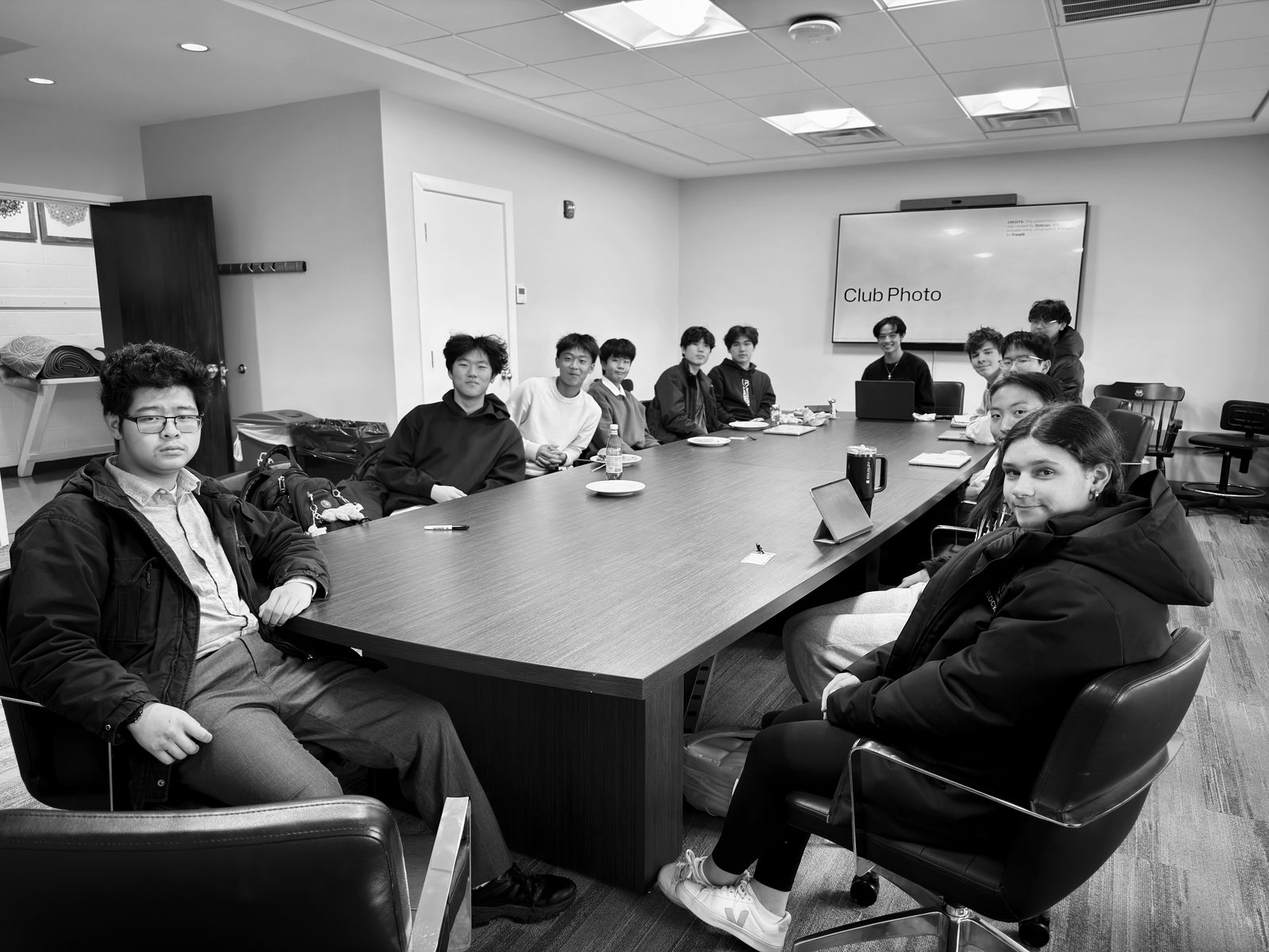 About a dozen students seated around a long dark conference table, turned toward the camera; a wall screen behind them reads “Club Photo”.