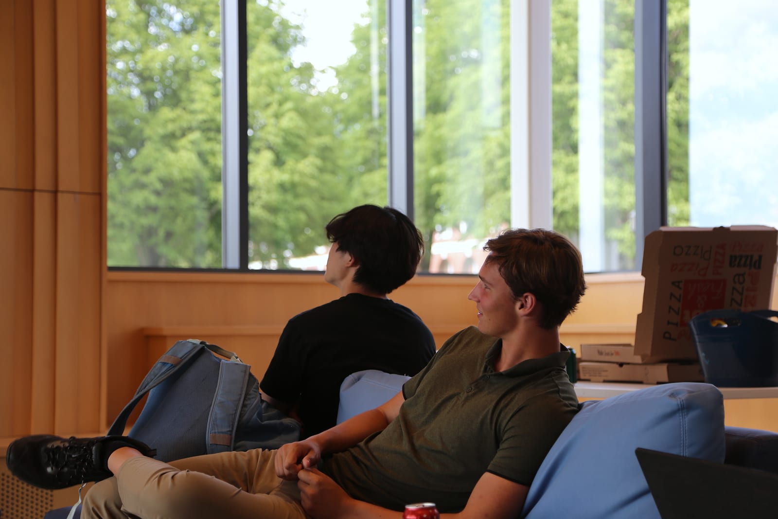 Two students on a sofa watch a talk off to the left; one smiles. Behind them, pizza boxes sit by the windows, with trees outside.
