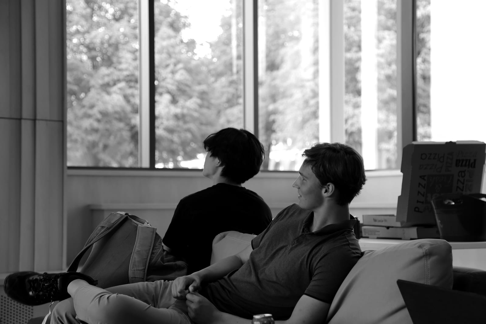 Two students on a sofa watch a talk off to the left; one smiles. Behind them, pizza boxes sit by the windows, with trees outside.