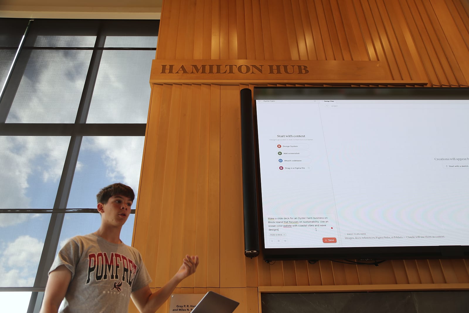 A student in a gray Pomfret T-shirt speaks and gestures below a “Hamilton Hub” sign. On the large screen beside them, a design tool shows a typed prompt asking for a slide deck for an oyster farm business on Block Island.
