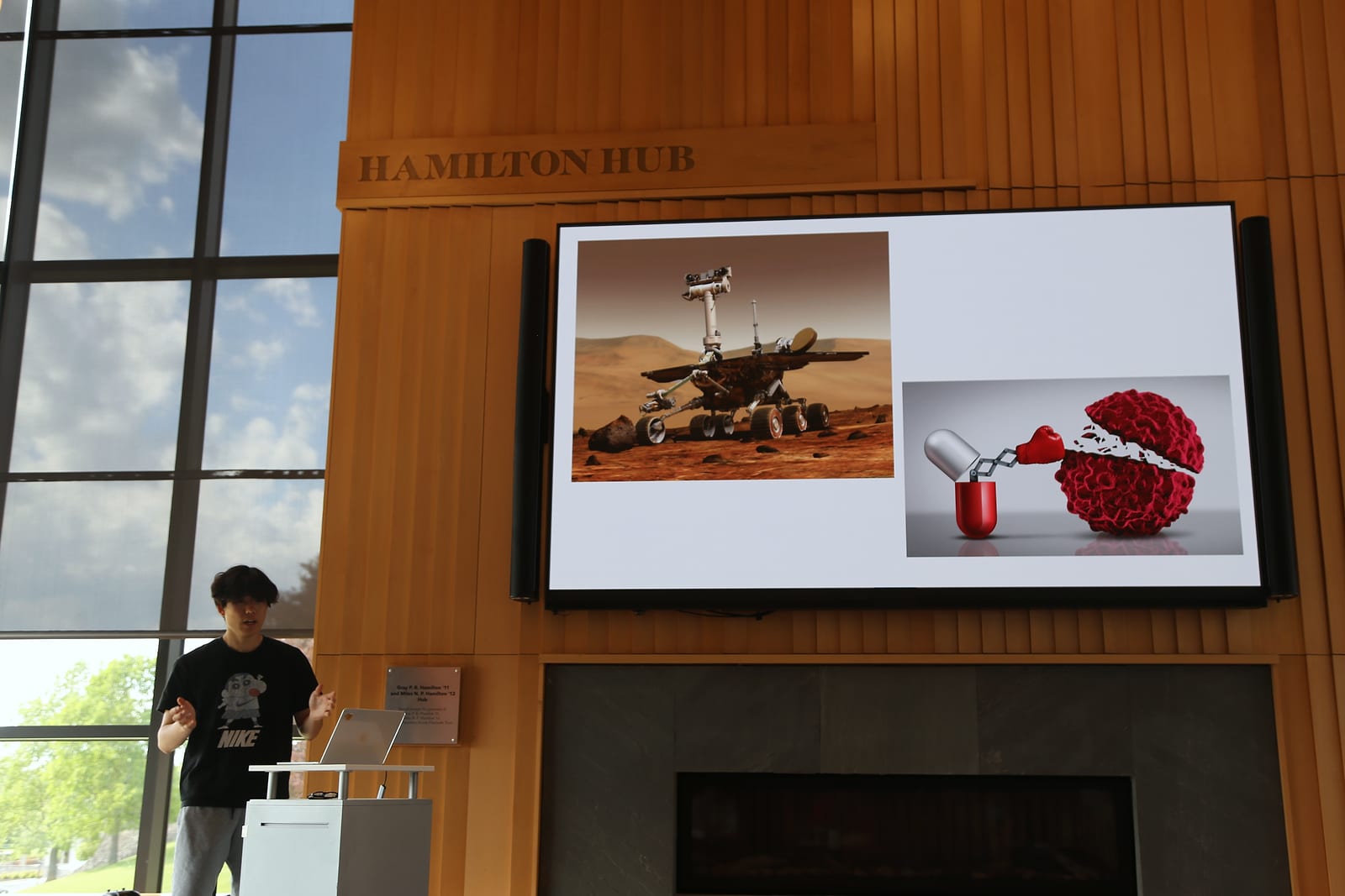 A student speaks from a lectern in the lower left corner. Above, a large screen under the “Hamilton Hub” sign shows two pictures: a rover on the surface of Mars, and a pill capsule with a boxing glove punching a cluster of cells.