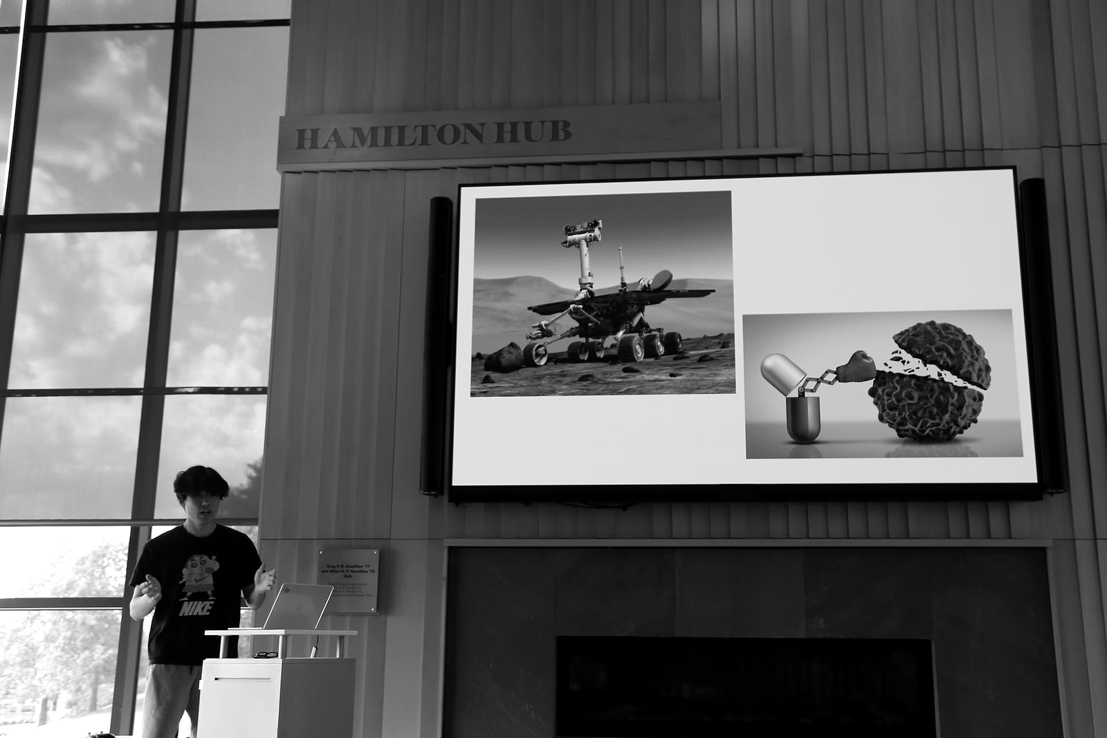 A student speaks from a lectern in the lower left corner. Above, a large screen under the “Hamilton Hub” sign shows two pictures: a rover on the surface of Mars, and a pill capsule with a boxing glove punching a cluster of cells.
