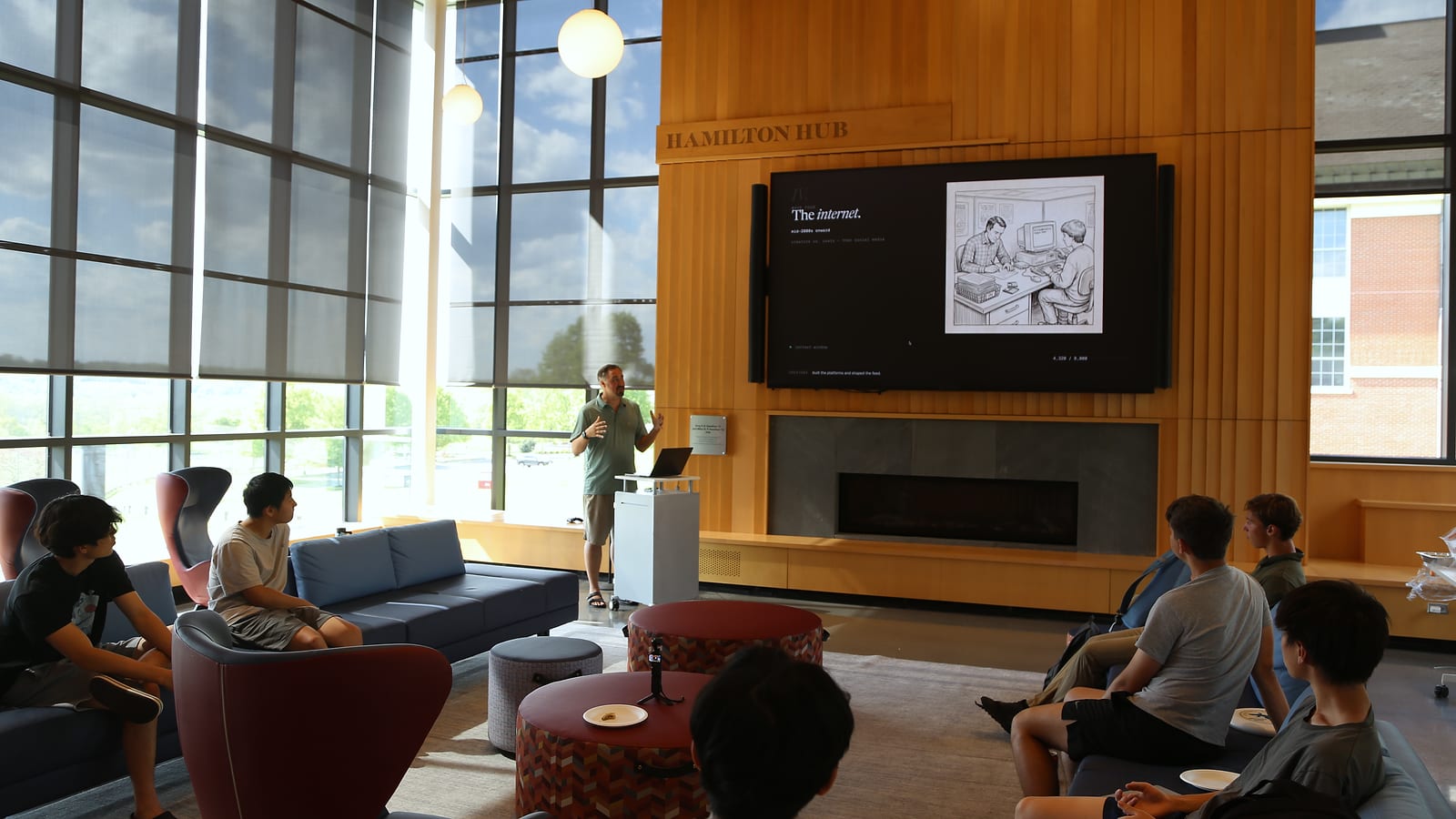A sunlit room with floor-to-ceiling windows and a tall wood-paneled wall under a “Hamilton Hub” sign. A presenter at a lectern speaks beside a large screen that reads “The internet.” while students listen from sofas and armchairs in the foreground.