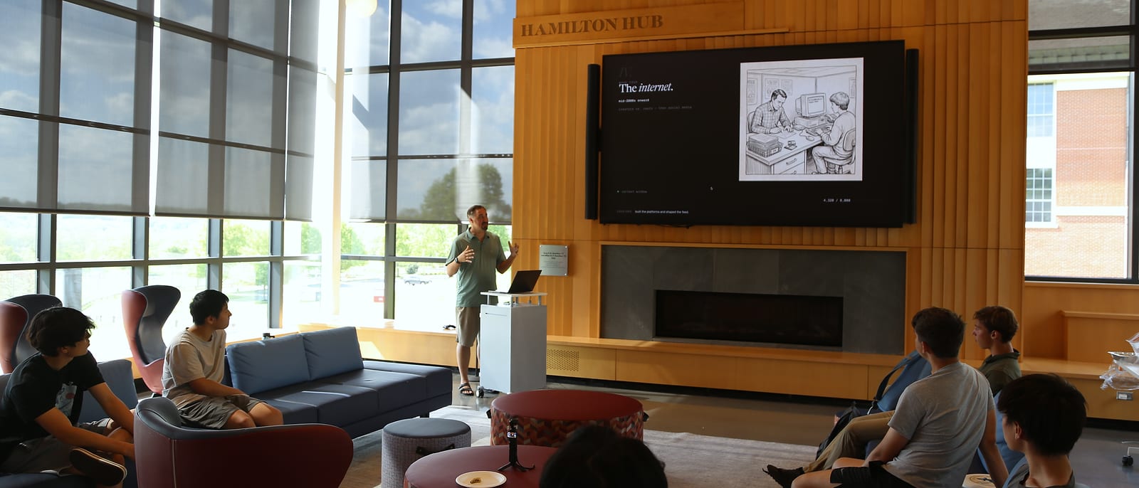 A sunlit room: tall windows on the left, a wood-paneled wall with a large screen that reads “The internet.”, a presenter at a lectern, and students on sofas watching.