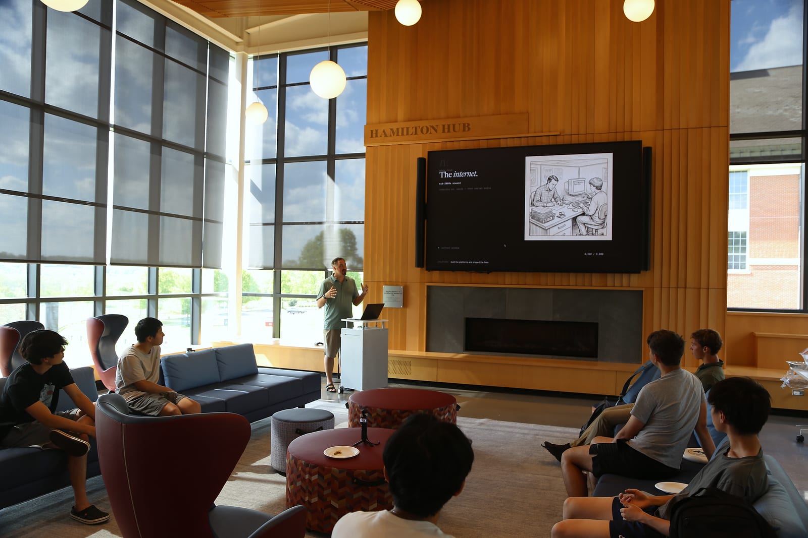A sunlit room with floor-to-ceiling windows and a tall wood-paneled wall under a “Hamilton Hub” sign. A presenter at a lectern speaks beside a large screen that reads “The internet.” while students listen from sofas and armchairs in the foreground.