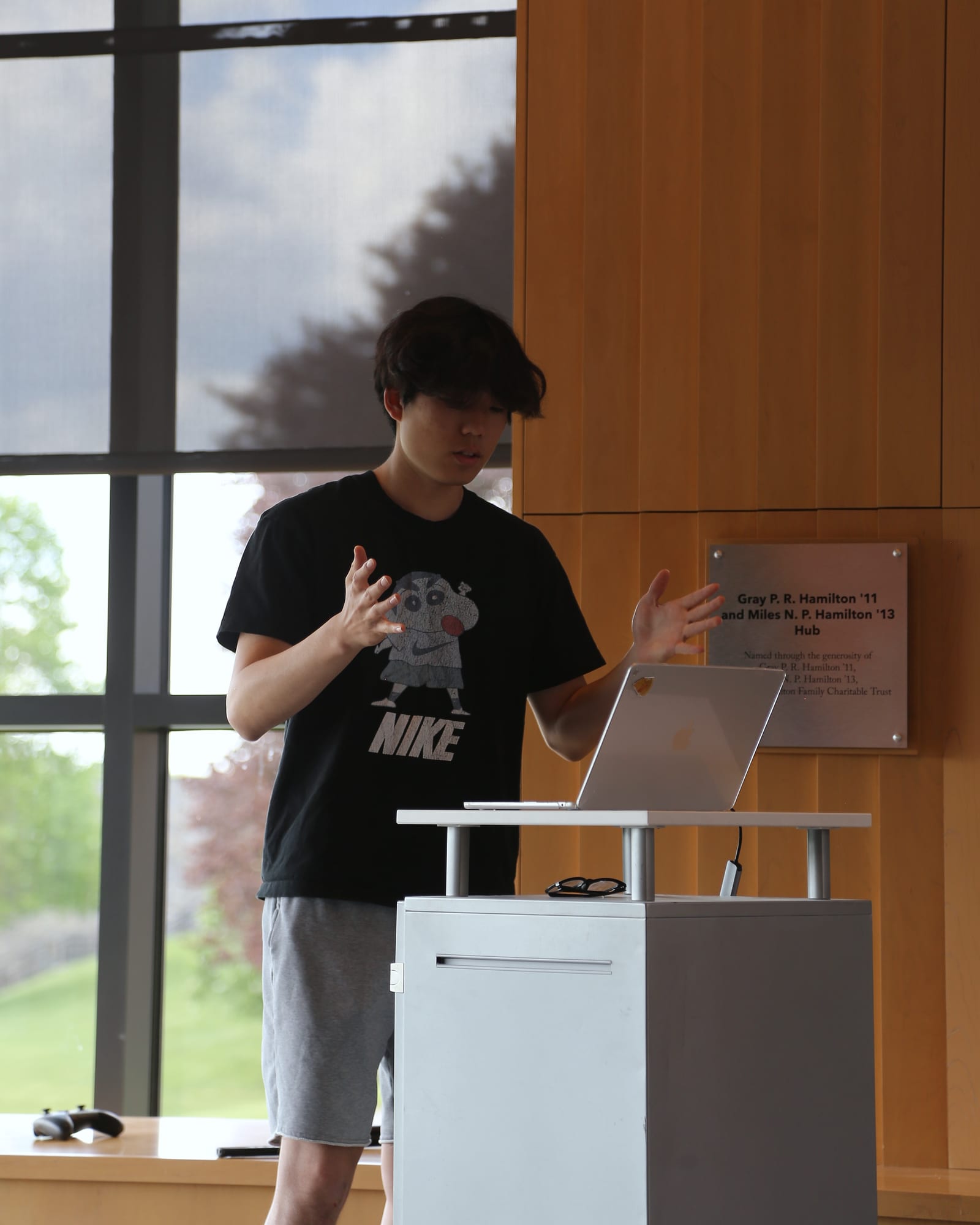 A student in a black T-shirt with a cartoon Nike print stands at a lectern, both hands raised mid-sentence, looking down at an open laptop. Tall windows and a wood-paneled wall behind.